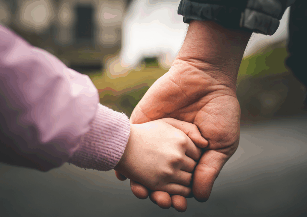 יד מבוגר גדולה אוחזת ברכות בידו הקטנה של ילד כסמל לביטחון, ליווי וקשר בין-דורי.